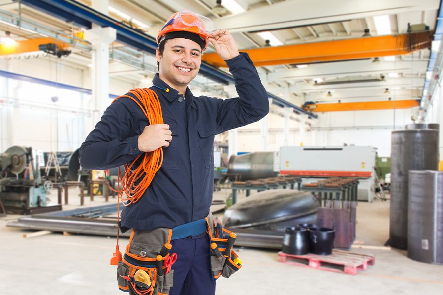 Contractor Insurance - Man Smiling in a Factory Warehouse While Holding Wiring Equipment and Wearing a Hardhat. Toolbelt and Other Protective Gear